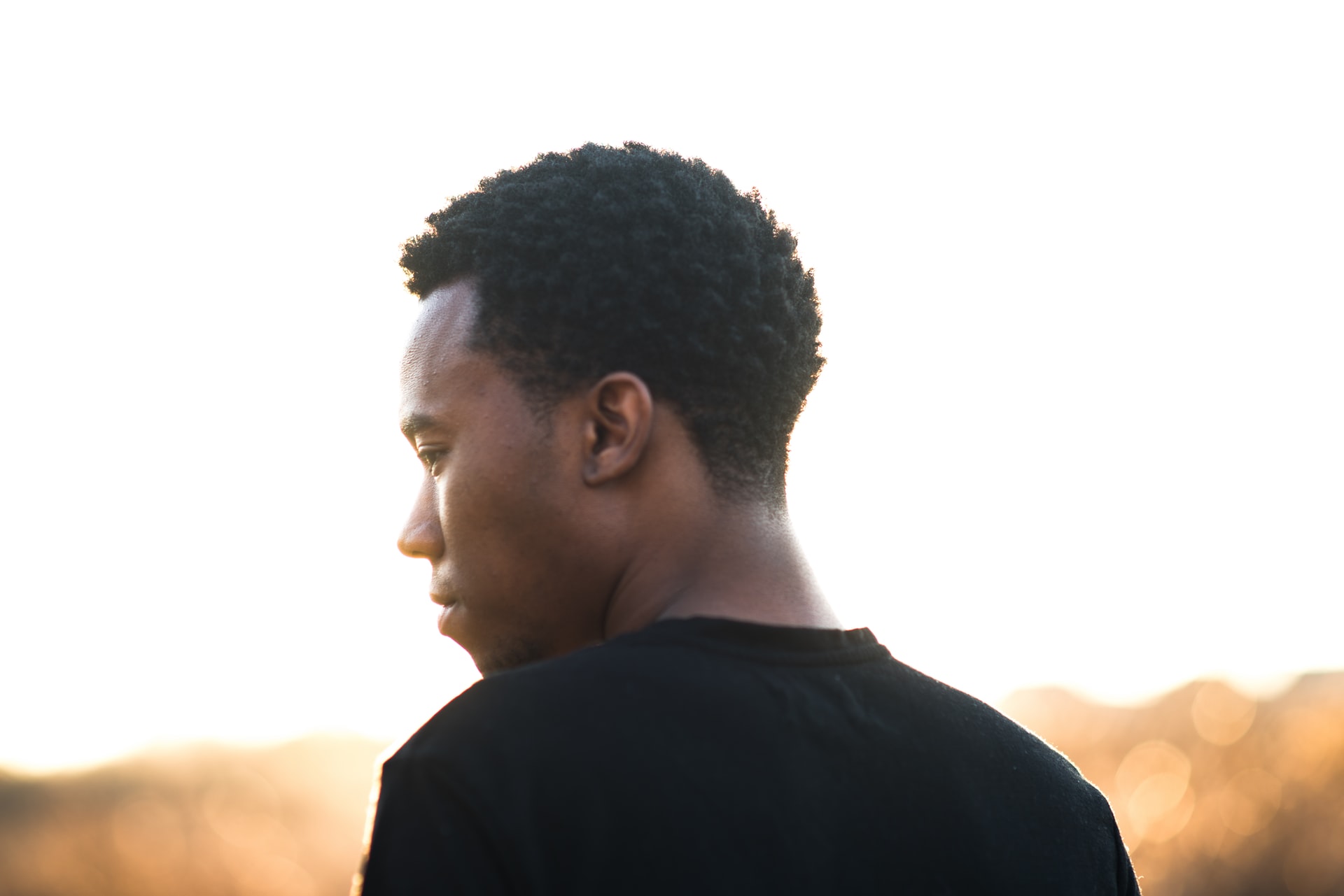 A young man wearing a black shirt standing in profile outside against an unfocused, bright background