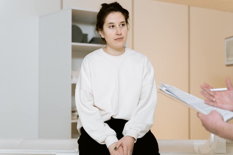 A young woman wearing a white sweater sitting on a medical exam table.