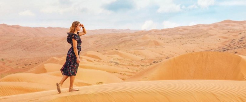 woman wearing floral dress blocking sun with hand while walking on sand dune in desert