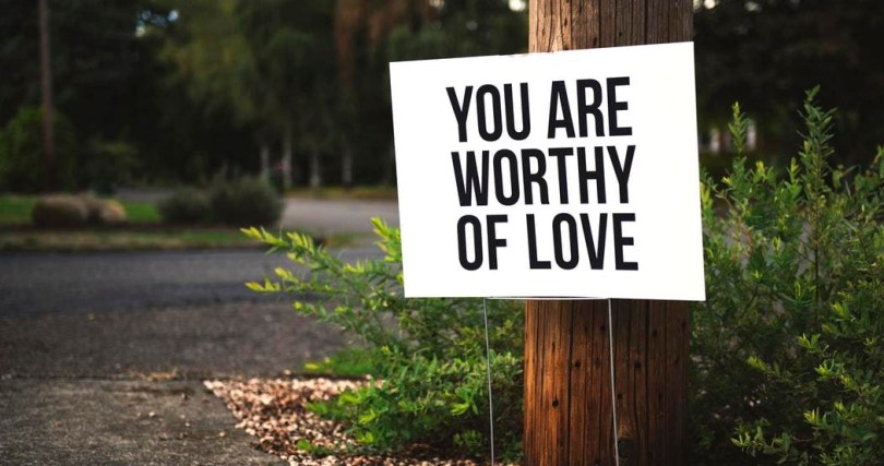 white sign with black capital letters reading "you are worthy of love" near telephone pole, green bush, and asphalt walkway