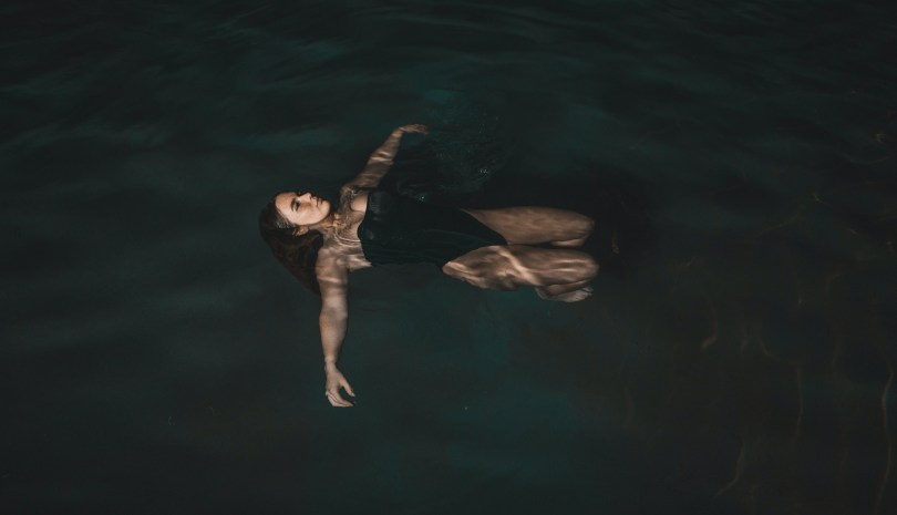 A woman wearing a black one-piece swimsuit floating in dark water with her knees bent and her arms outstretched to the sides