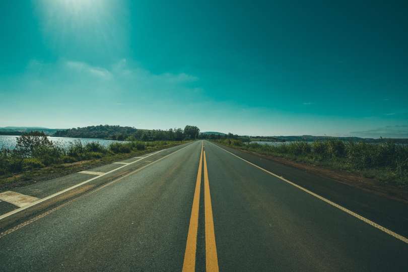 A stretch of straight open road with a body of water to the left and blue sky above