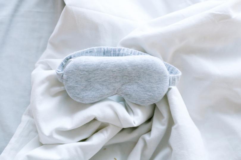 A padded grey sleep mask with an elastic band arranged on white linens