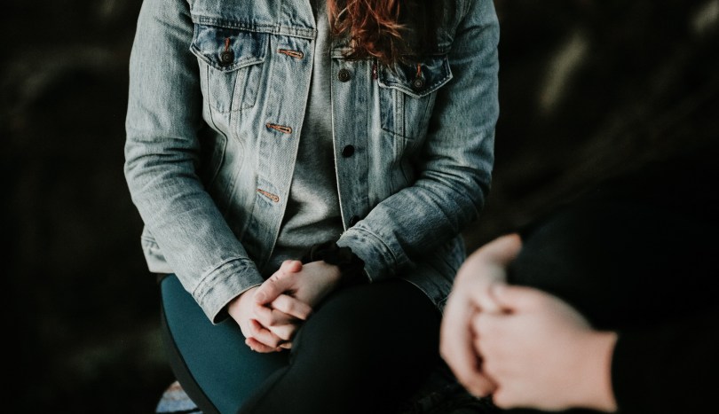 An unidentifiable woman wearing a jean jacket sitting with her hands clasped in her lap and her legs crossed while talking to a therapist in the foreground.