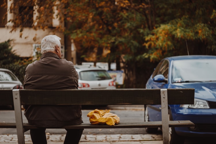 older-man-with-white-hair-sitting-on-park-bench-with-back-toward-camera-and-yellow-garment-folded-on-bench-beside-him-and-blue-car-parked-ahead