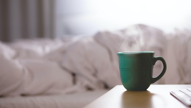 green-mug-with-steam-rising-sitting-on-side-table-with-rumpled-sheets-on-bed-in-background