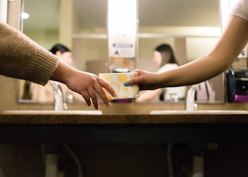 Two women in a public bathroom passing a pad in a yellow and white wrapper between them