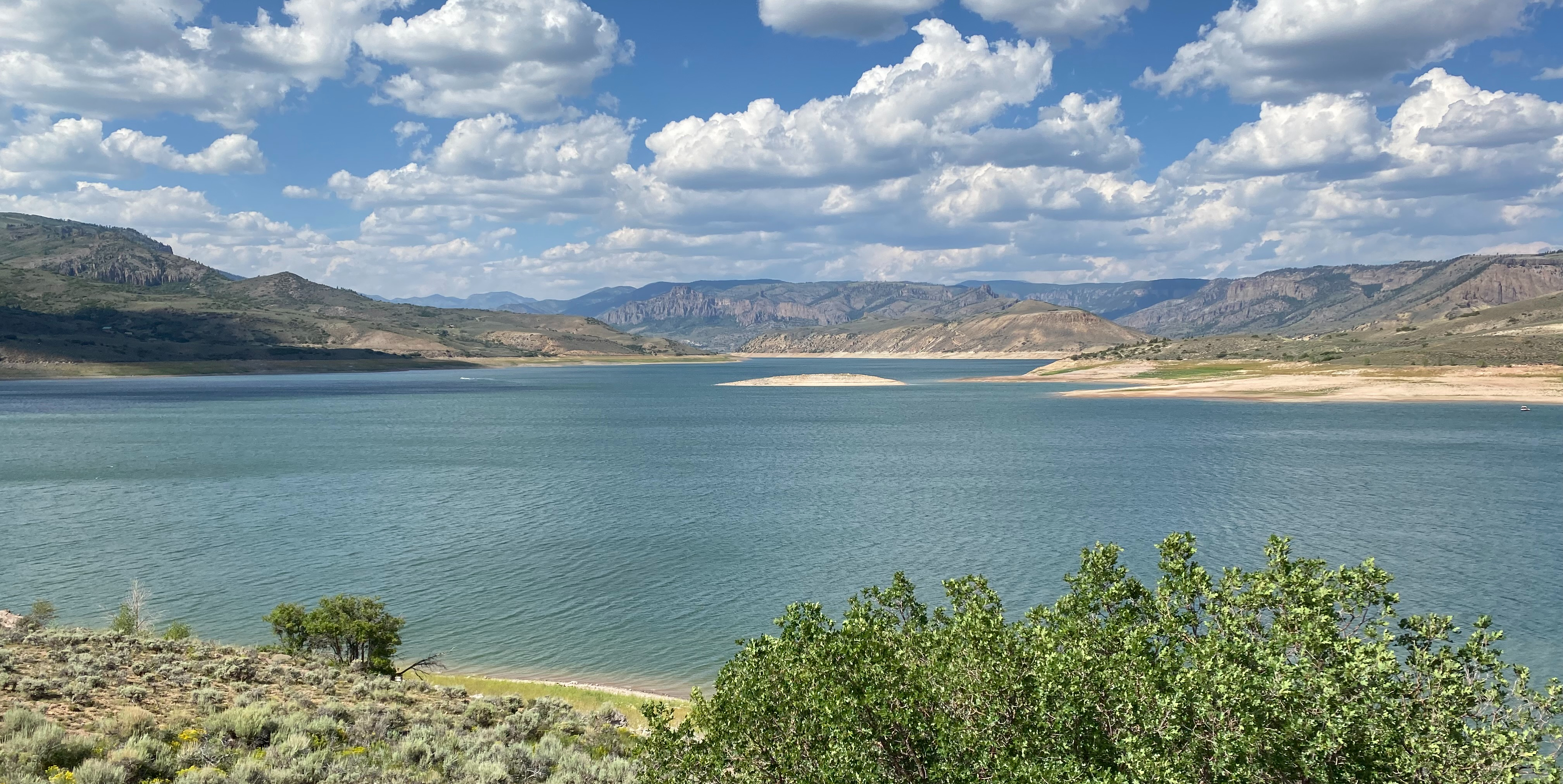 A sunny landscape with a large blue lake, green vegetation, mountains, and a blue sky with fluffy white clouds