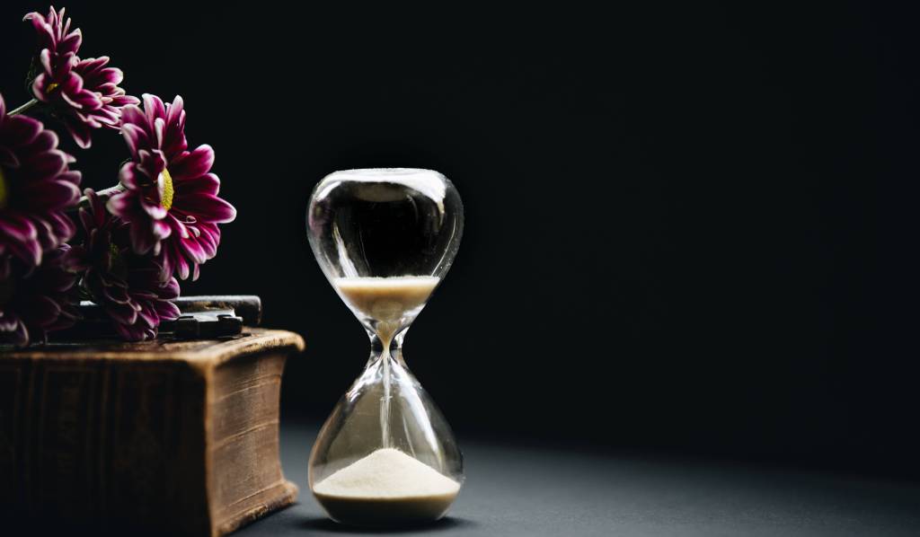 An hourglass with sand falling from one side to the other sitting next to an old book with purple flowers resting on top. Dramatic lighting with a dark background.