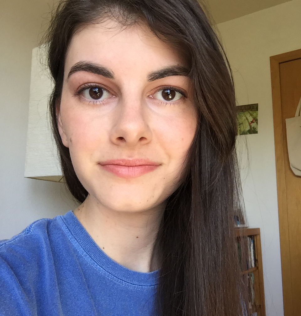 A young woman with straight, brown hair draped over one shoulder is looking at the camera and smiling gently.