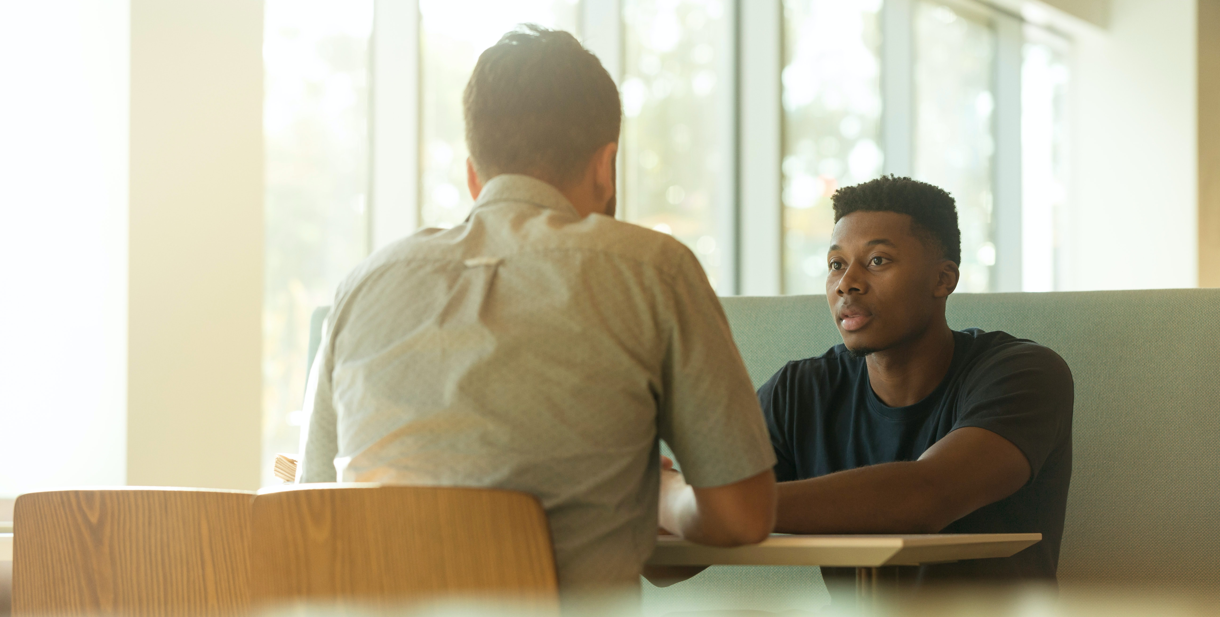 Two men facing each other while seated at a table in a sunny lobby having a discussion.